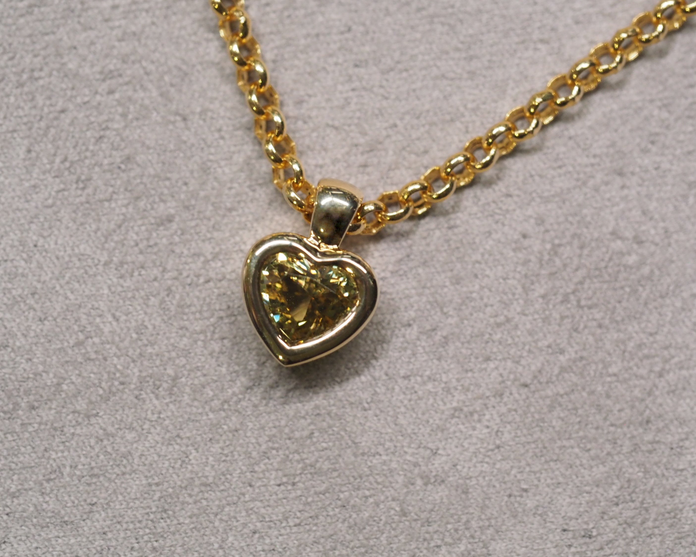 A gold heart-shaped pendant necklace with a yellow-green heart-shaped gemstone, resting on a textured gray background.