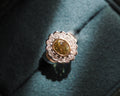 A close-up shot of a vintage-style ring featuring a scalloped silver-toned setting with a central round brown gemstone and a halo of small clear stones, resting on a dark teal velvet ring box.