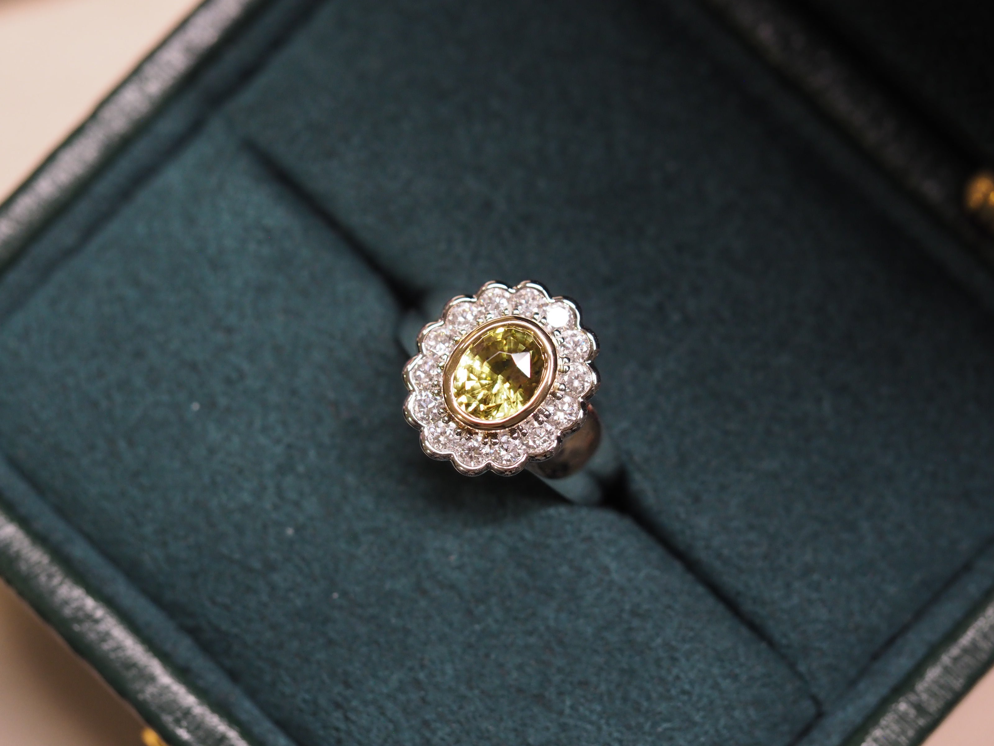 A yellow chrysoberyl ring with a halo of small clear stones, displayed in a dark green velvet ring box.