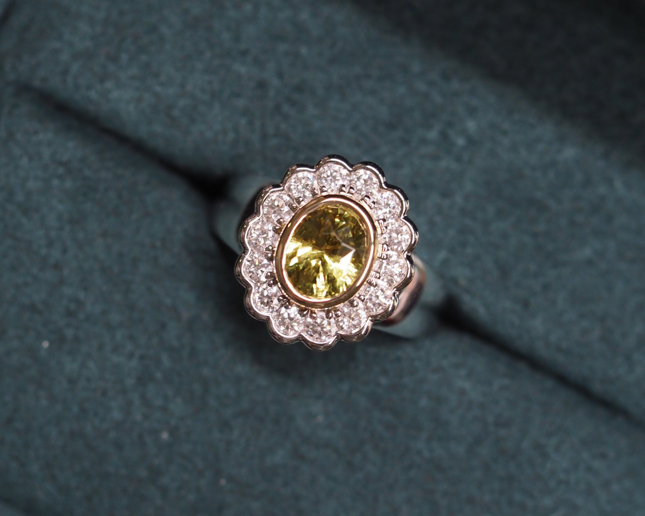 A two tone gold ring with a scalloped halo of small clear stones and a central oval yellow chrysoberyl, displayed on a dark textured velvet surface.