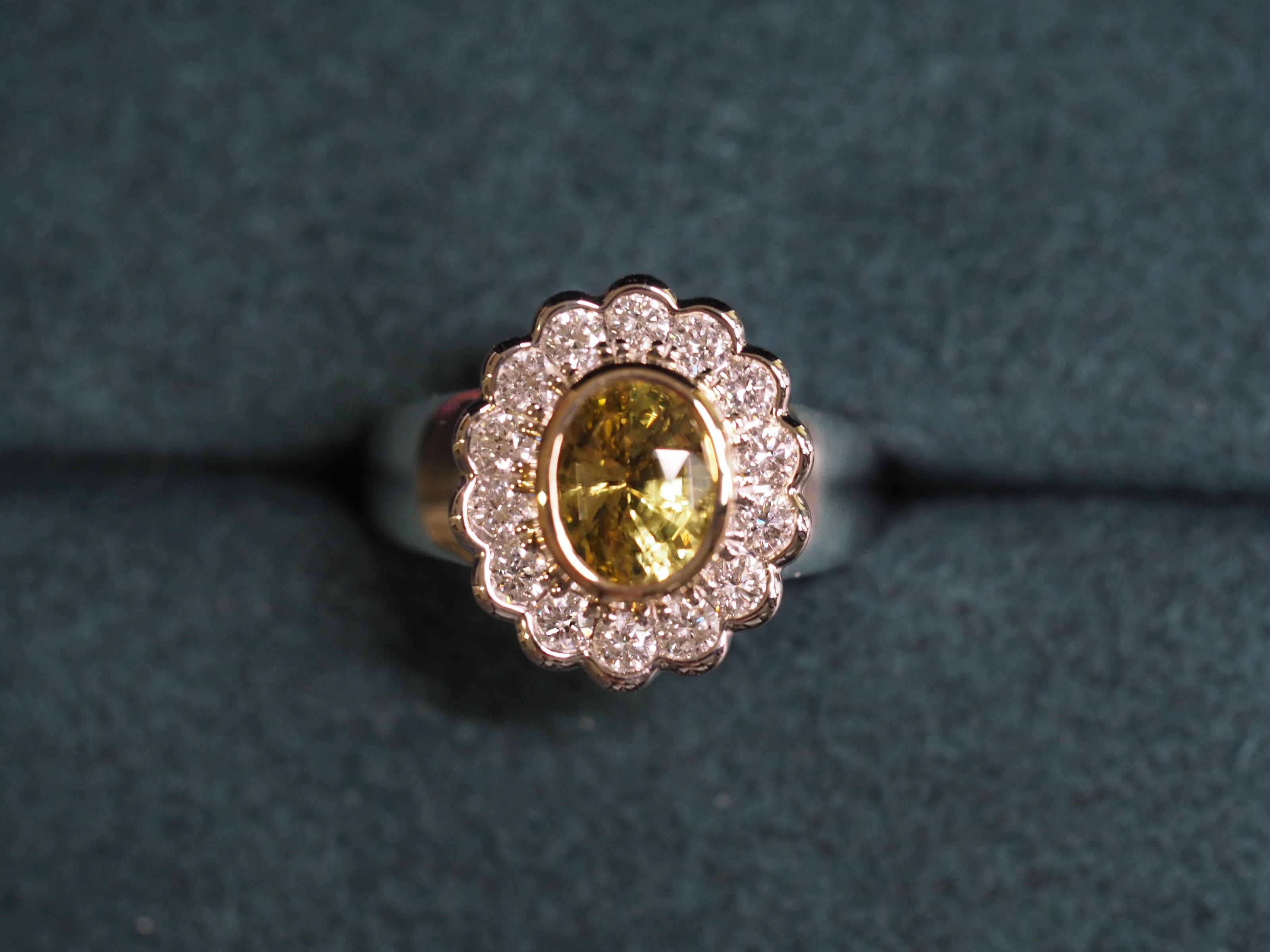 A two tone gold ring featuring an oval yellow chrysoberyl surrounded by a halo of small clear stones, displayed on a dark textured background.