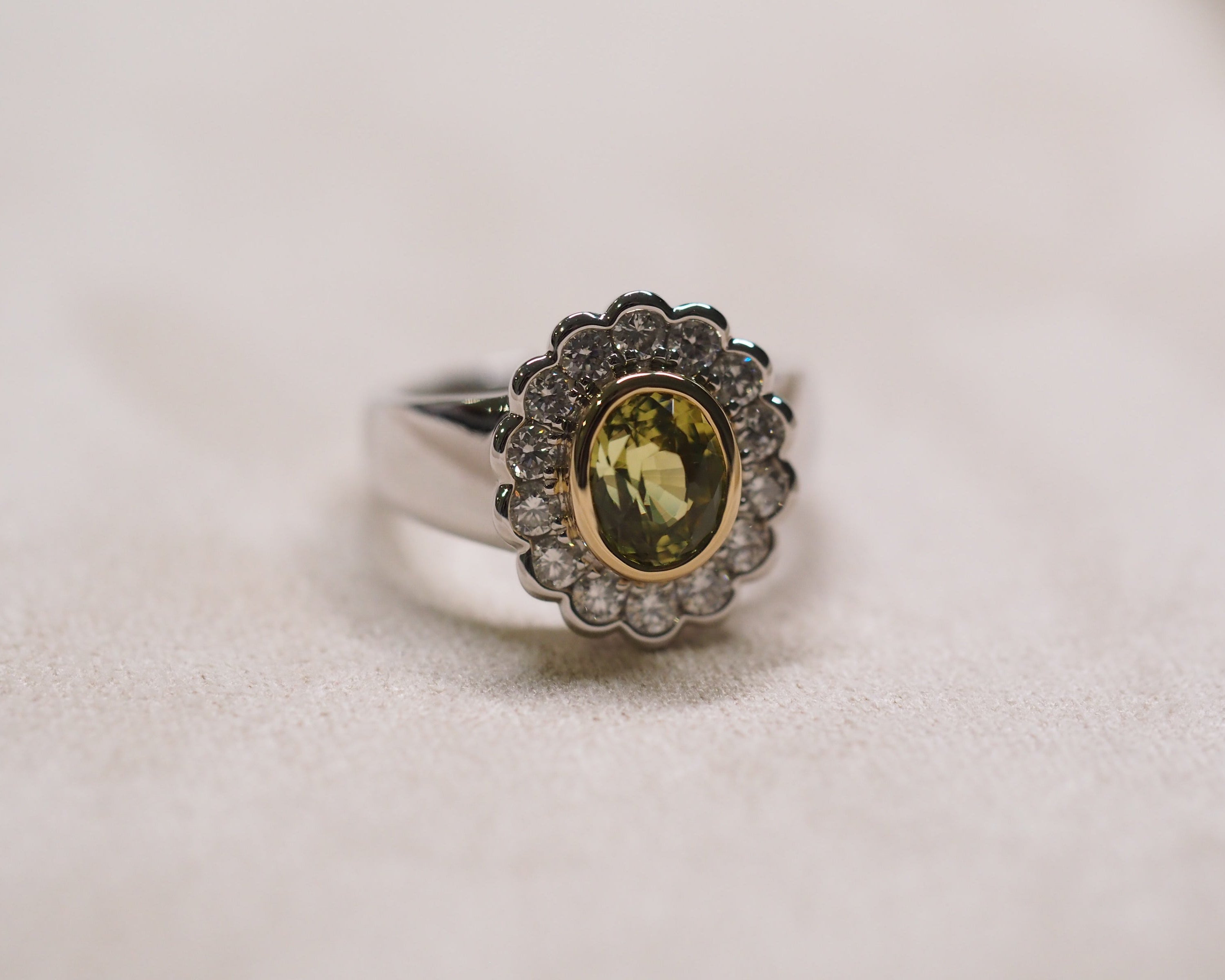 A vintage-style ring featuring an oval yellow-green center stone surrounded by a scalloped halo of small clear stones, set in a silver-toned band with a gold-toned inner ring, resting on a textured beige surface.