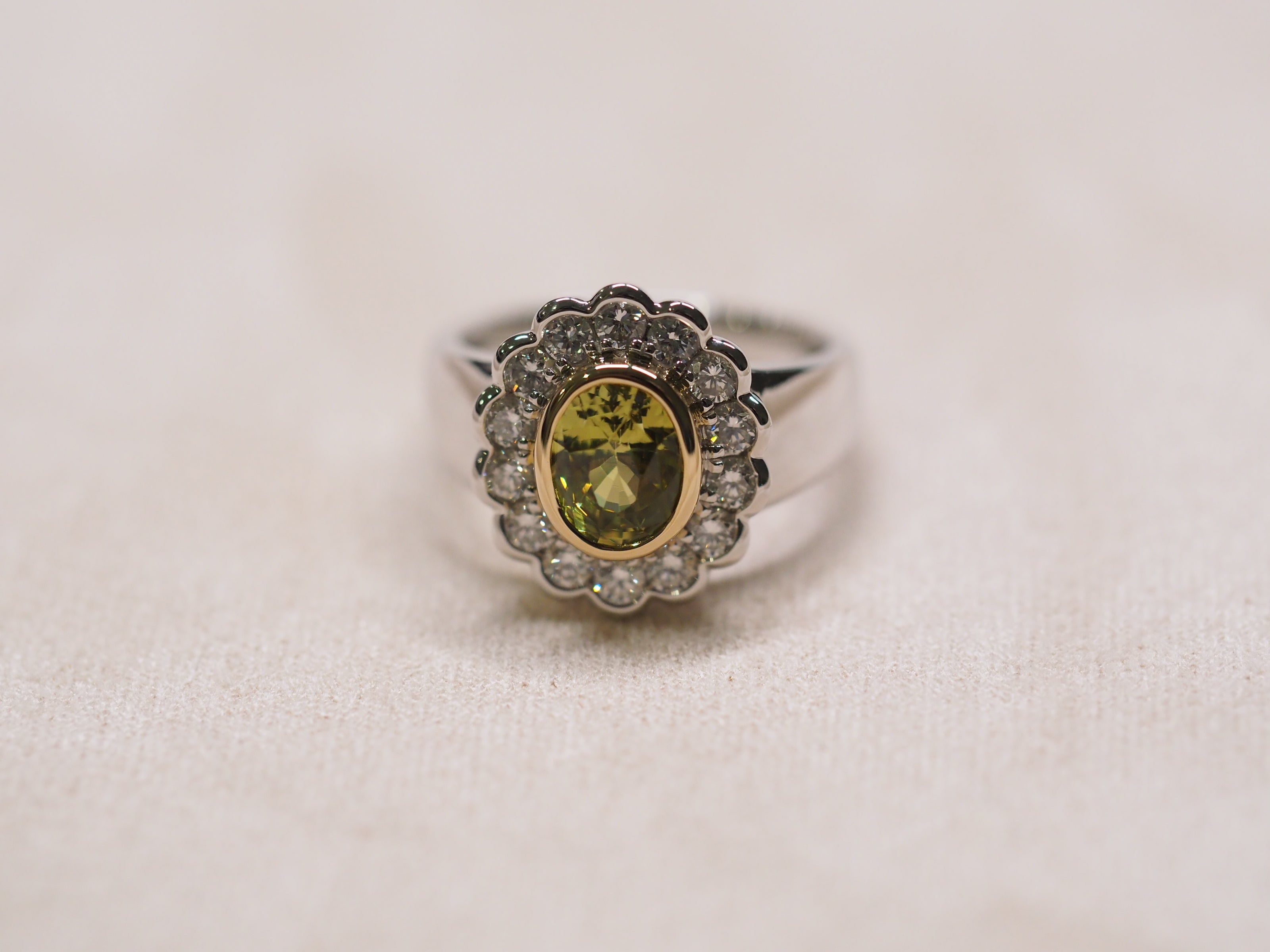 A silver ring featuring a central oval yellow-green gemstone surrounded by a double halo of small clear stones, with a gold-toned inner bezel, set against a light beige textured background.