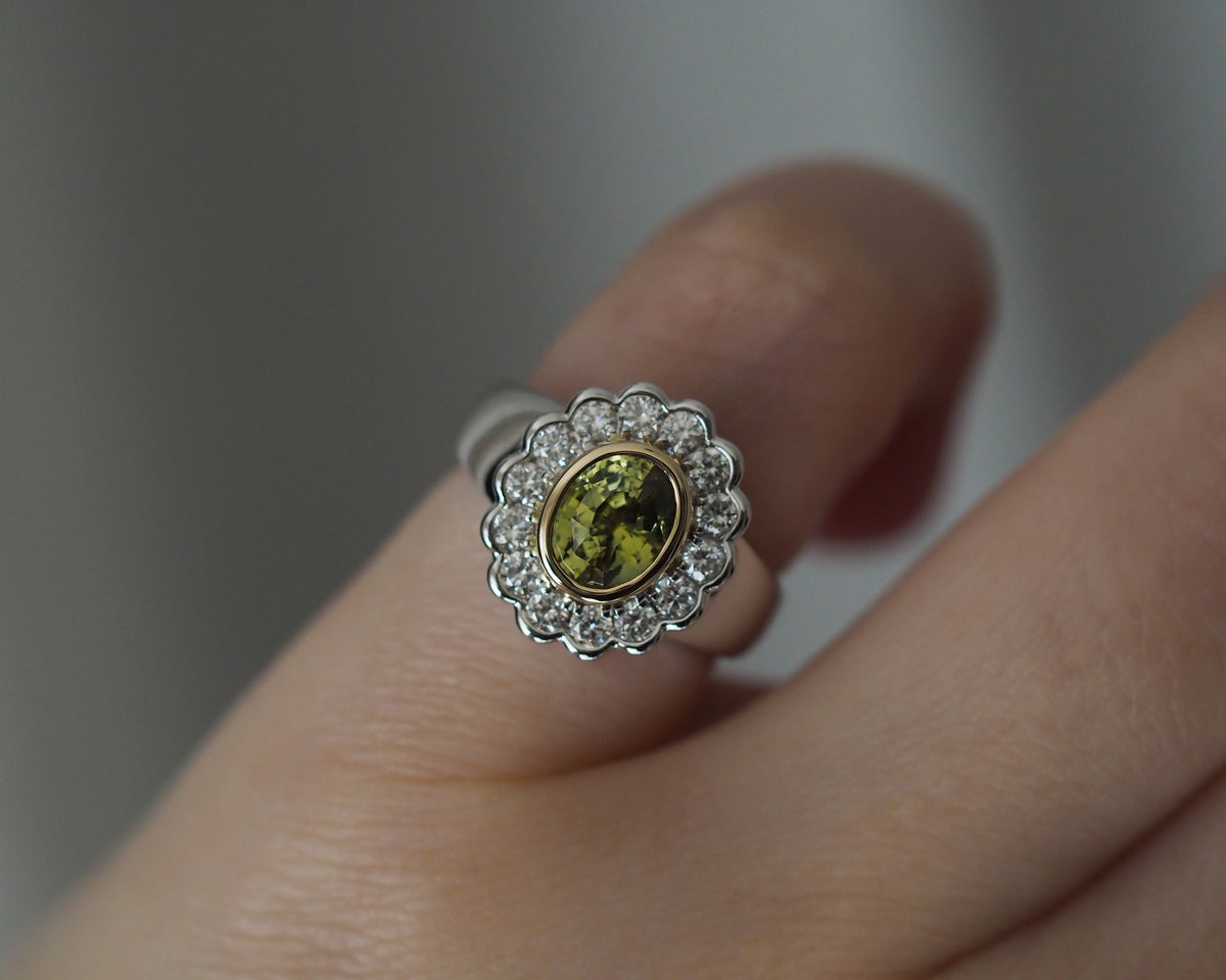 A close-up of a silver ring with a scalloped diamond halo and a central olive-green oval stone, worn on a finger.