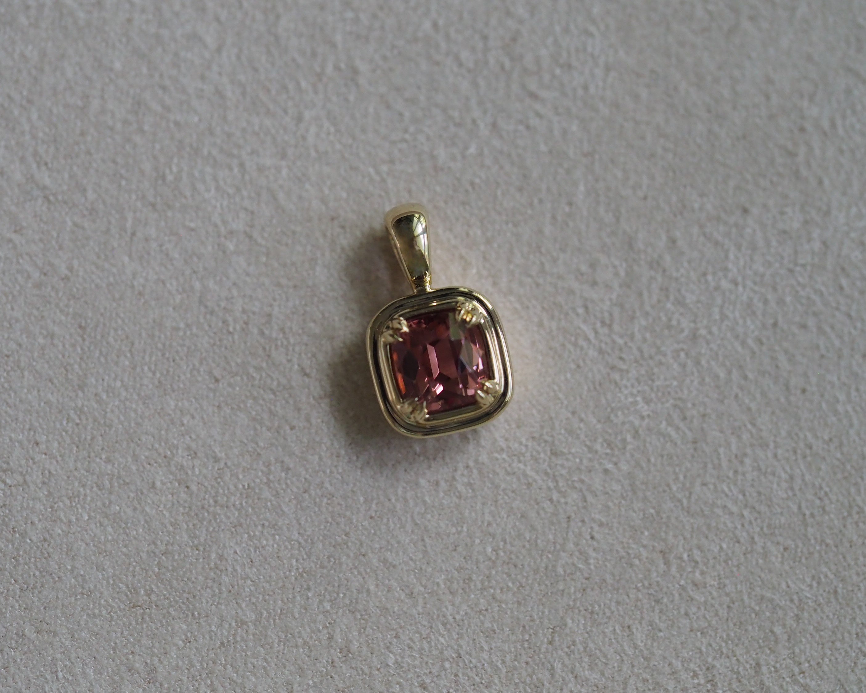 A gold pendant featuring a cushion-cut orangey pink gemstone, photographed on a textured light gray background.