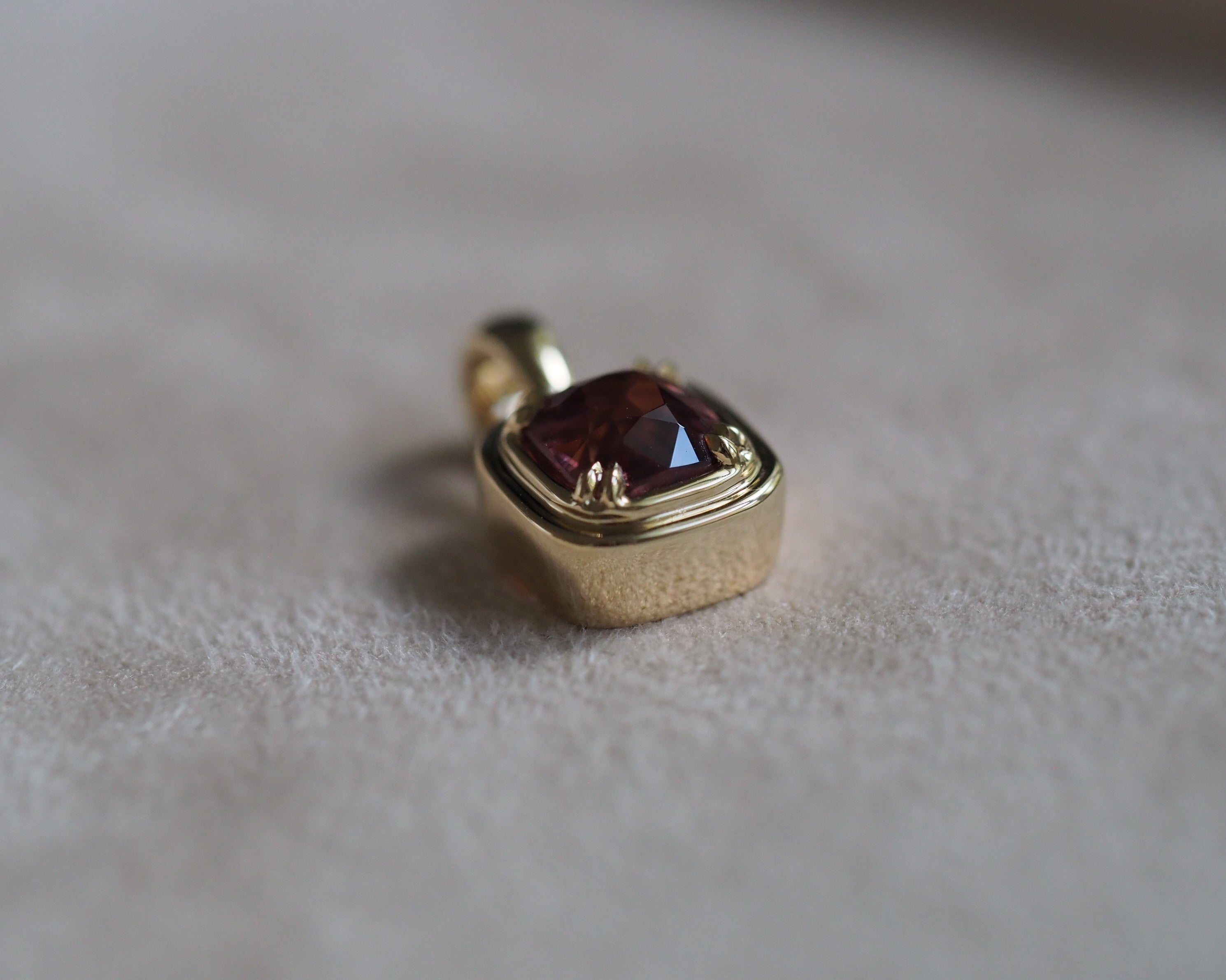 A gold pendant with a cushion-cut dark red gemstone, resting on a soft, textured beige fabric surface.