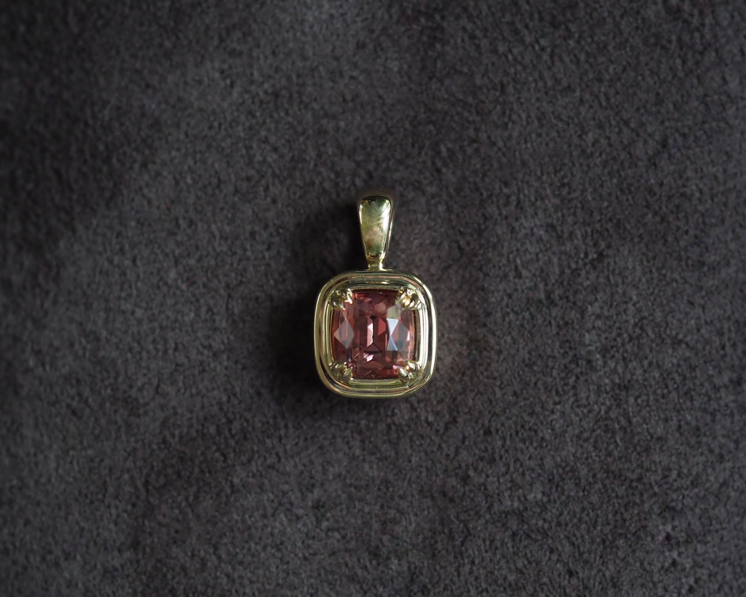 A square pendant featuring a cushion-cut pink gemstone in a gold bezel setting, displayed on a dark textured background.