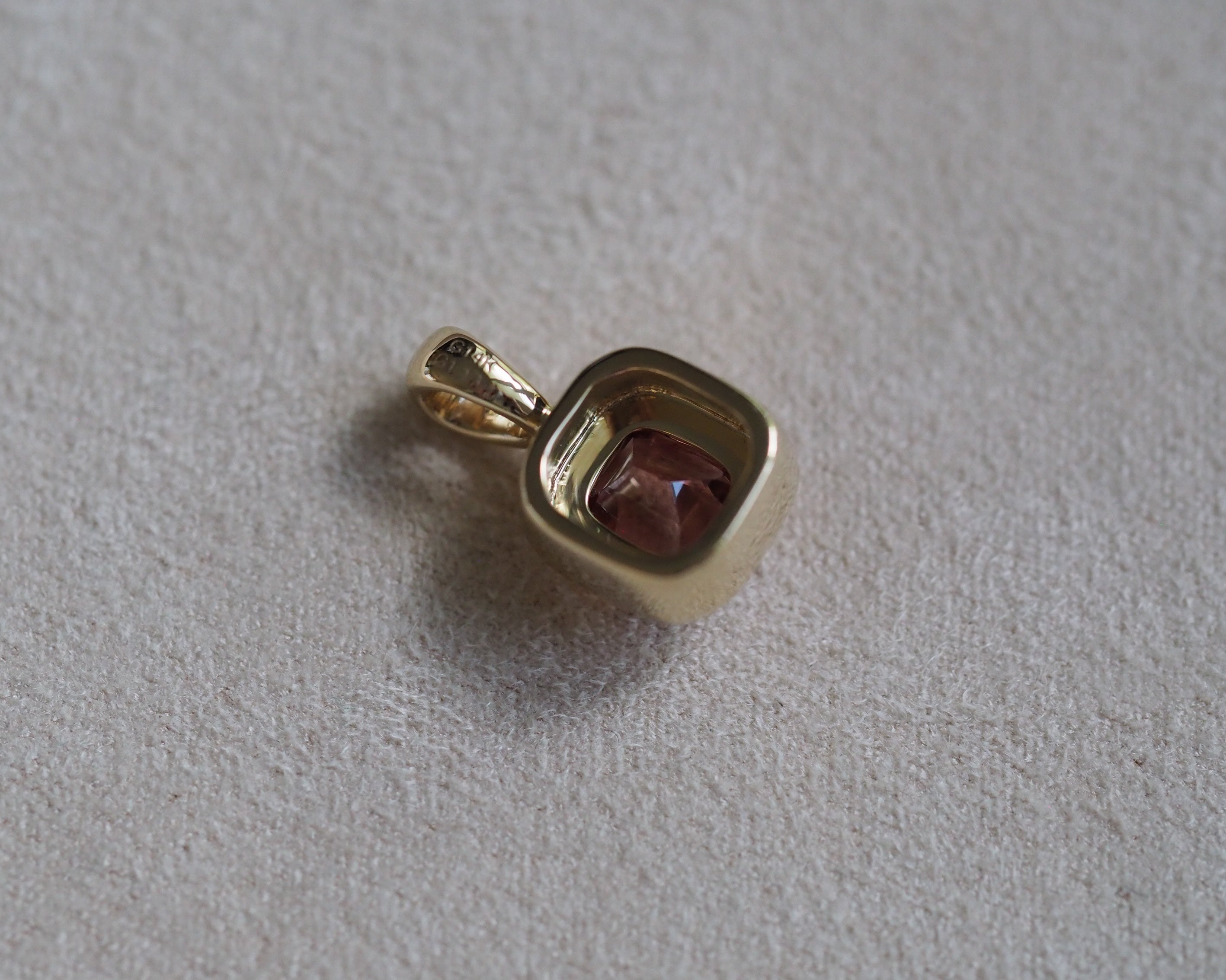 A small gold pendant with a square bezel setting holding a faceted reddish-brown gemstone, shown on a textured light gray surface.