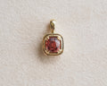 A gold pendant featuring a cushion-cut, faceted reddish-pink Malaya garnet, displayed on a textured beige surface.