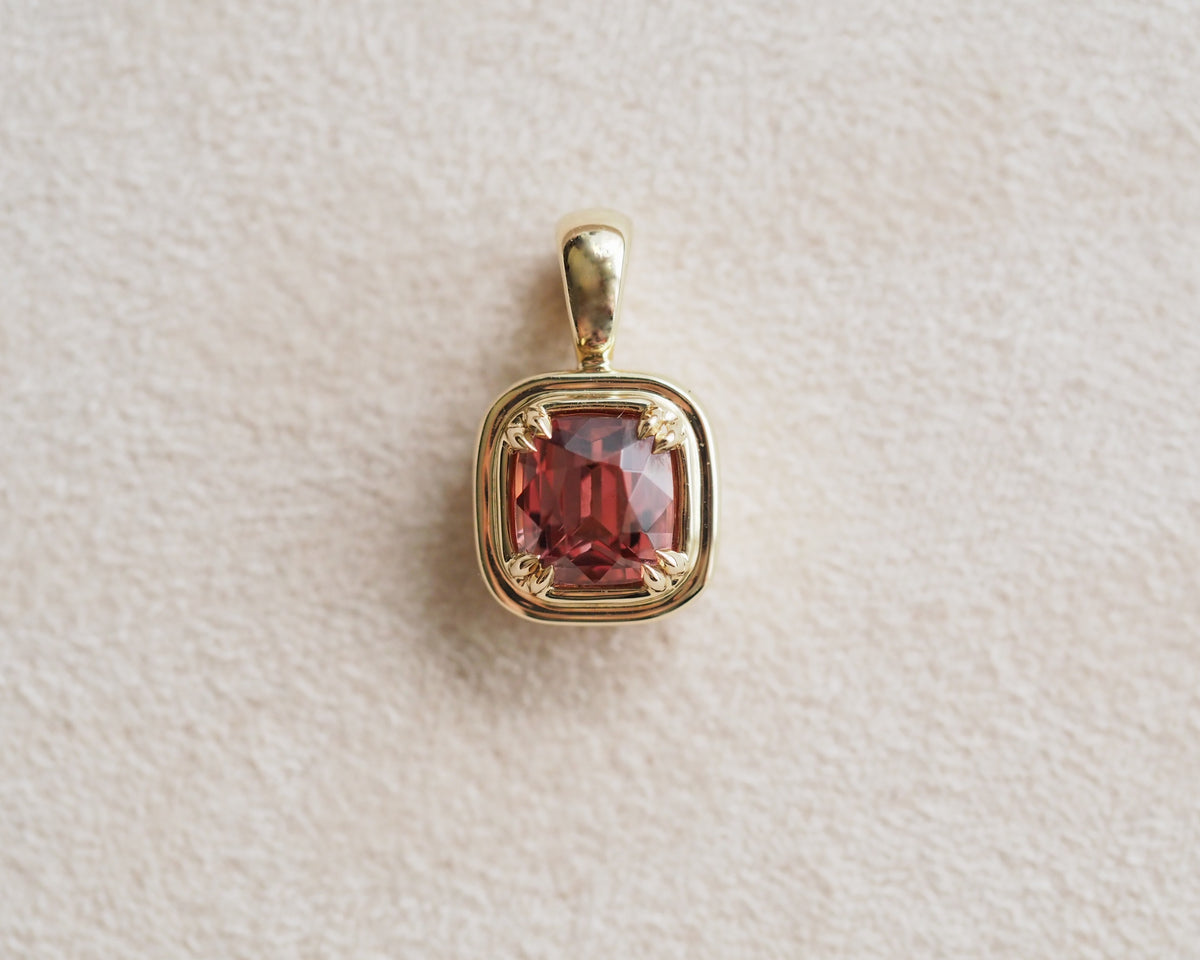 A gold pendant featuring a cushion-cut, faceted reddish-pink Malaya garnet, displayed on a textured beige surface.