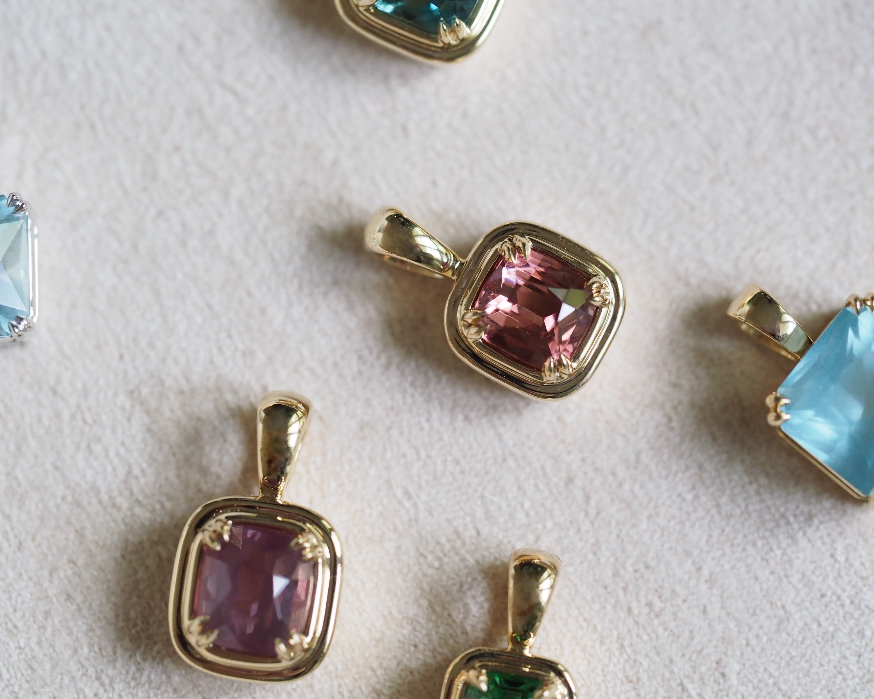 Gold square bezel pendants with colorful gemstones, including pink, purple, green, and blue, arranged on a textured white surface.