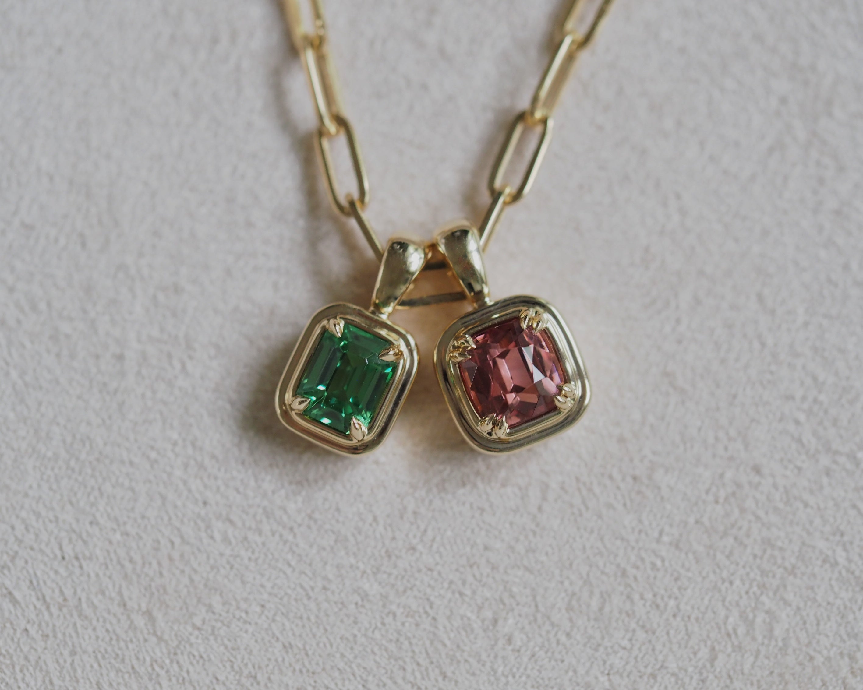 A close-up of a gold chain necklace with Robyn Tsavorite pendant and Juno Malaya Garnet pendant settings on a textured light gray background. One pendant features a green tsavorite, and the other features a orangey pink Malaya garnet, both set in yellow gold.