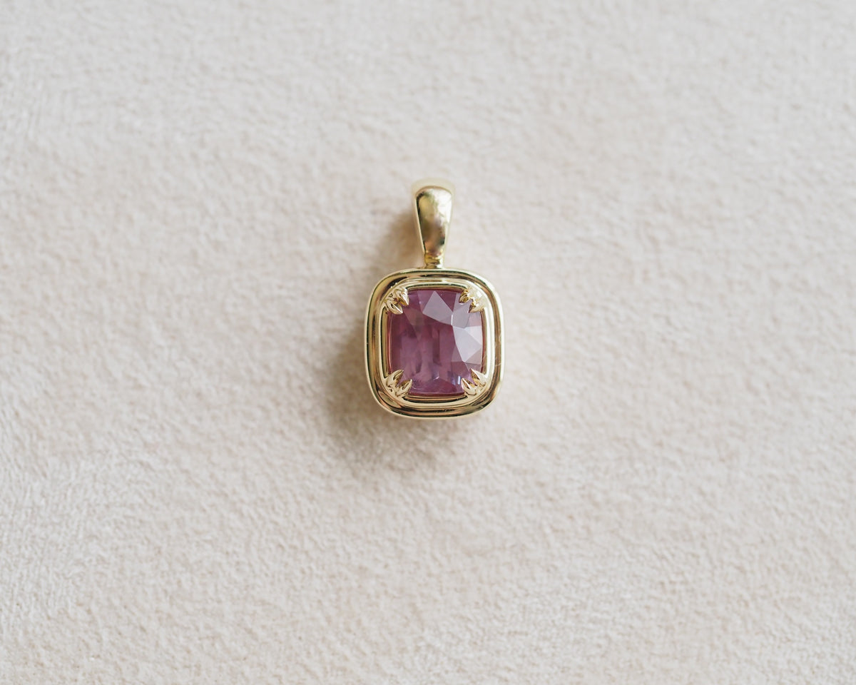A gold pendant featuring a square-cut, cushion-shaped pinkish-purple garnet, displayed on a textured off-white background.