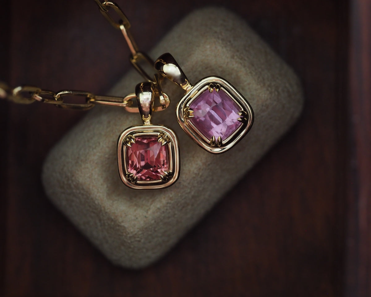 Two gold pendant necklaces with cushion-cut pink gemstones, one resting on a textured gray stone and the other on a dark wooden surface.
