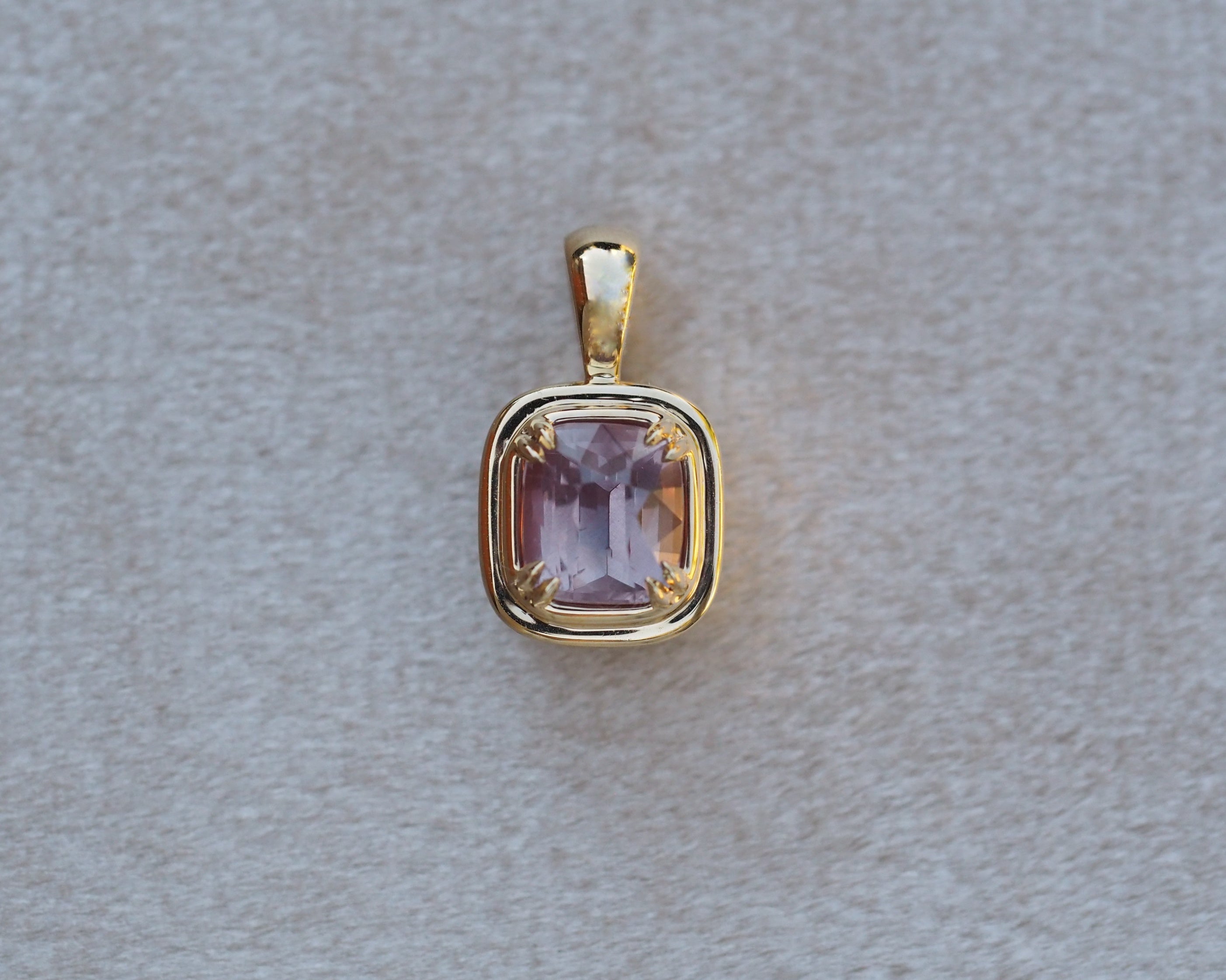 A gold pendant with a cushion-cut light purple gemstone, displayed on a textured light gray background.