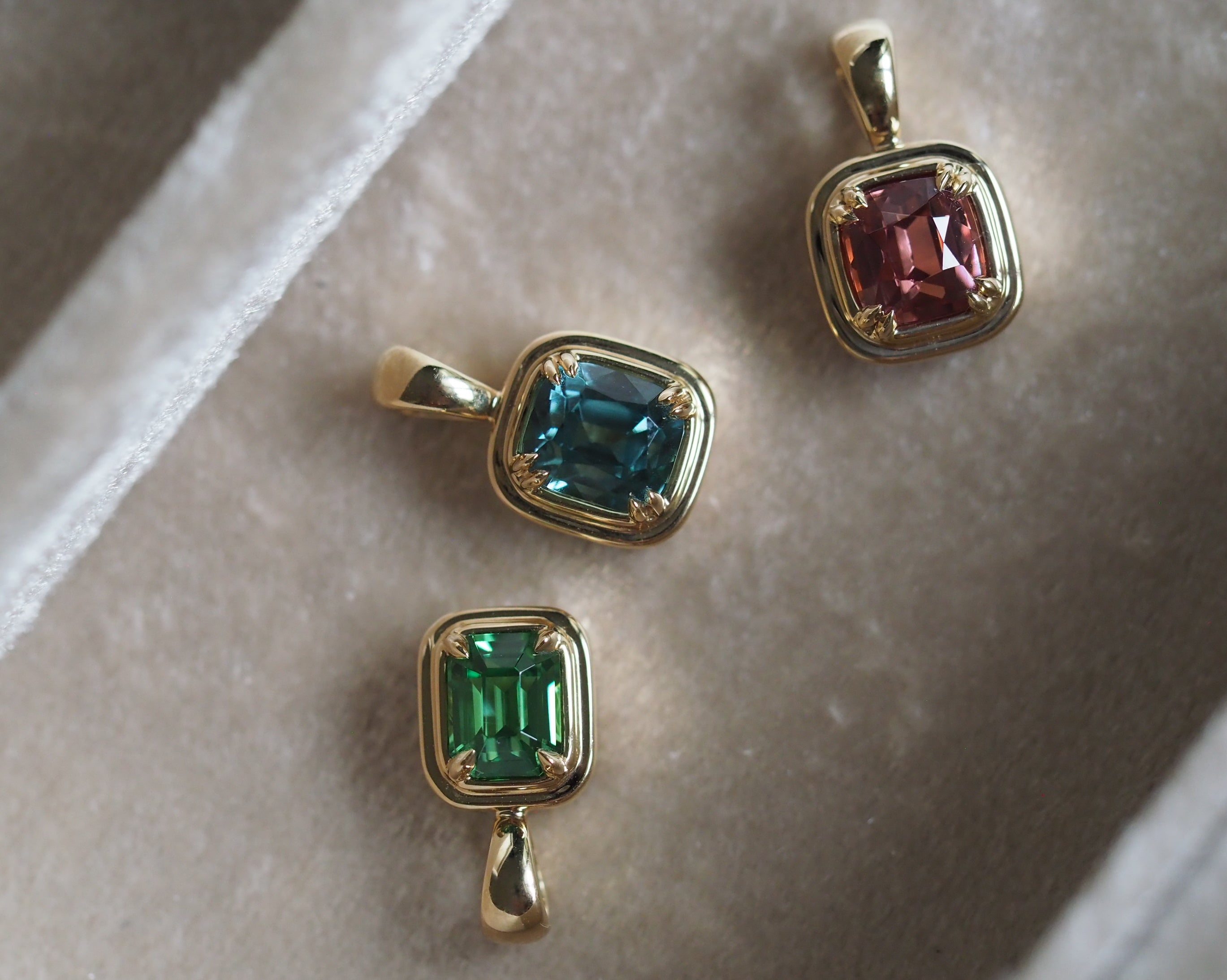 Three gold pendant charms with cushion-cut gemstones in green, blue, and pink, displayed on a beige velvet jewelry box.