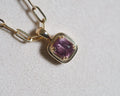 A gold pendant necklace featuring a cushion-cut milky pink gemstone in a gold stepped bezel setting, matched with a gold paperclip chain on a textured light gray background.