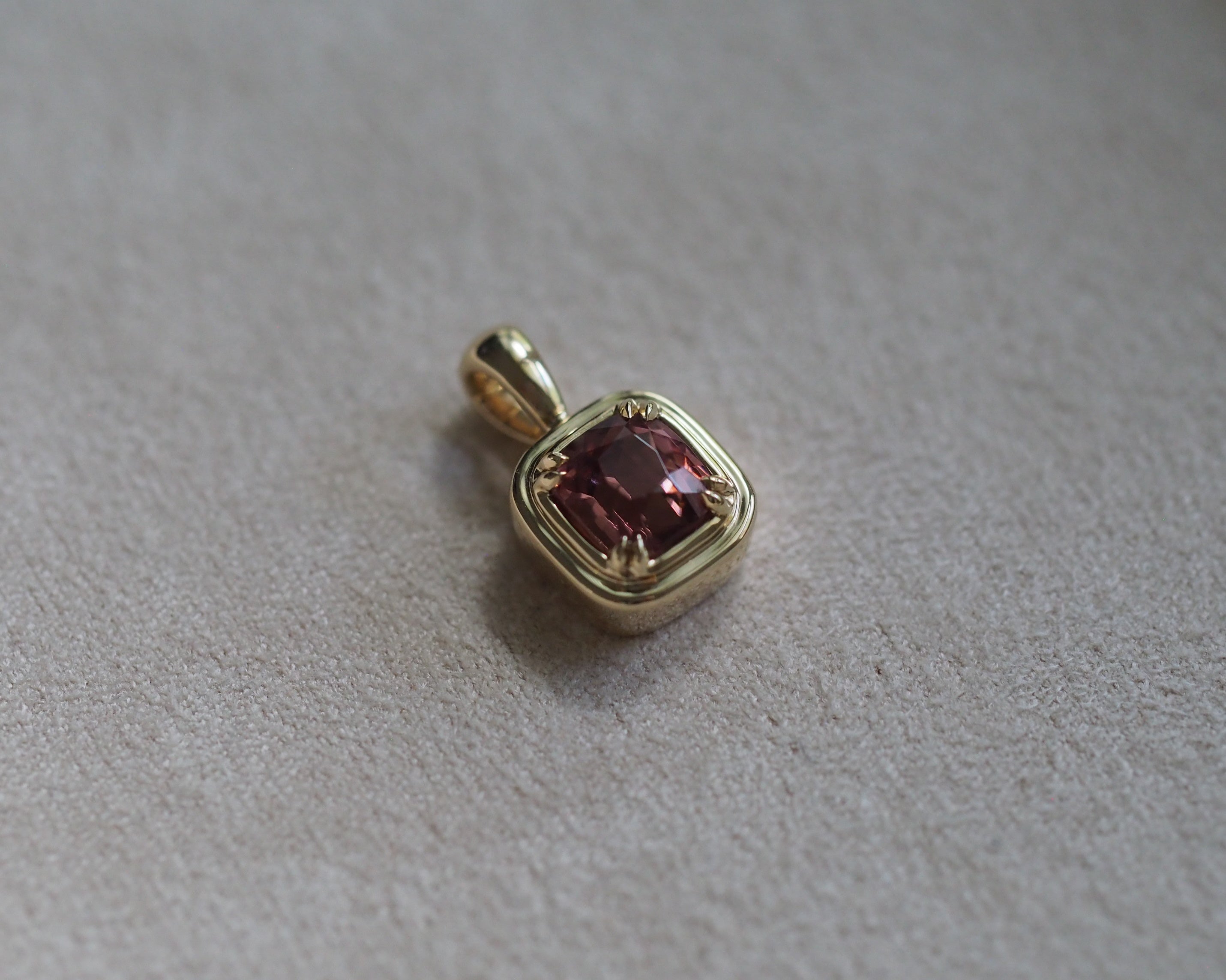 A small gold pendant with a square-cut orangey red gemstone, resting on a textured light gray surface.