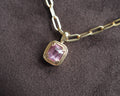 A gold chain necklace with a cushion-cut pink milky garnet pendant, displayed on a textured dark gray fabric background.