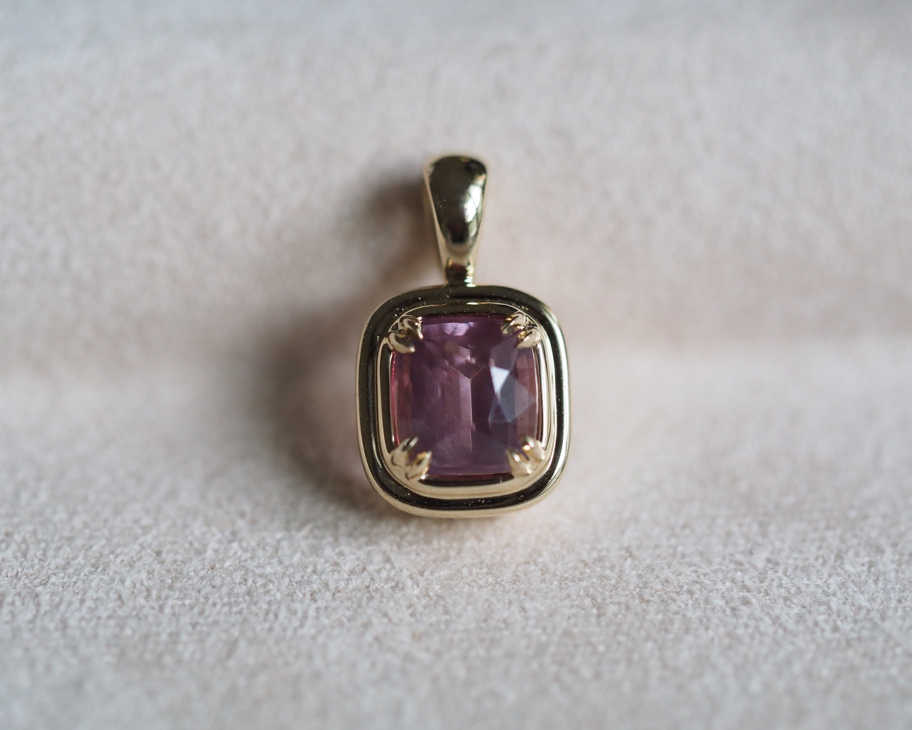 A gold pendant featuring a cushion-cut pink garnet in a four double prong setting, resting on a textured light beige surface.