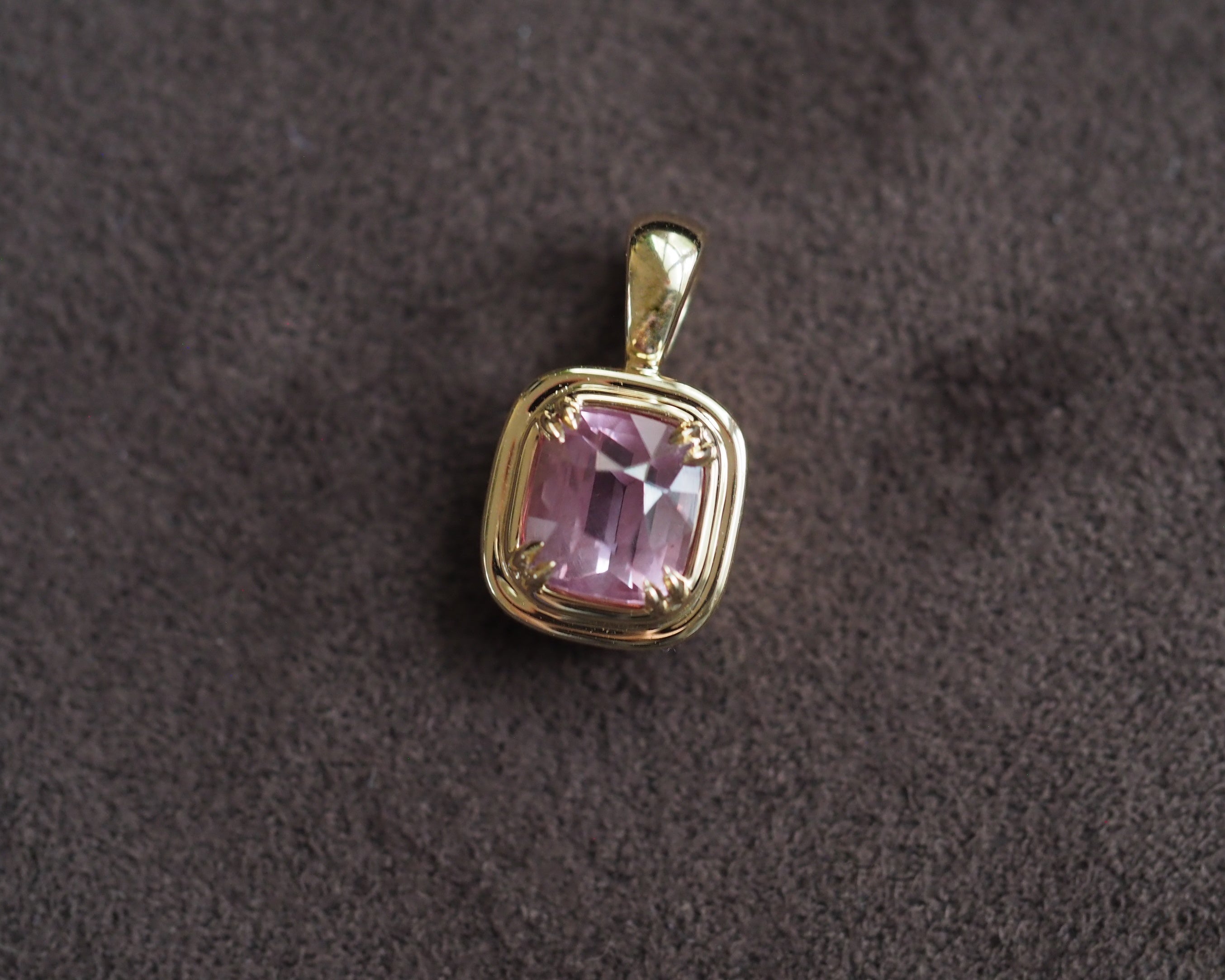 A gold pendant with a cushion-cut pink milky garnet, displayed on a textured dark brown surface.