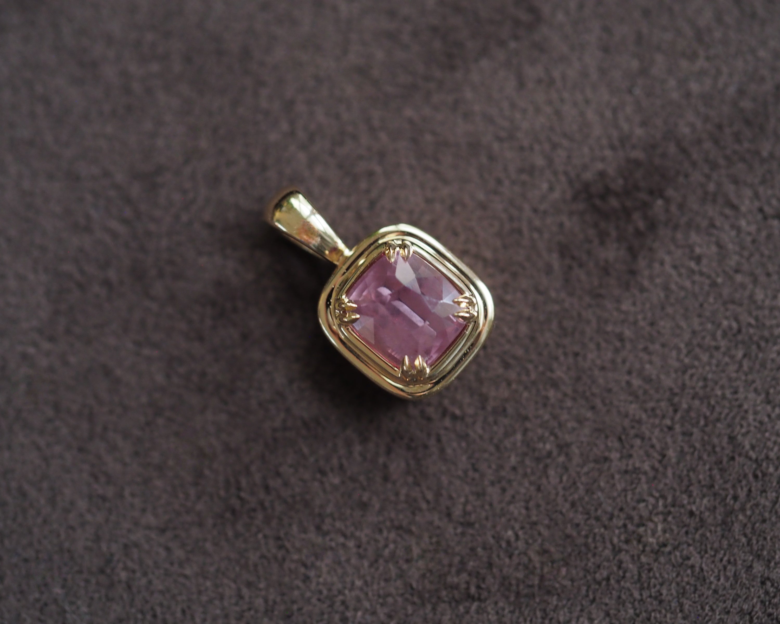 A gold pendant featuring a cushion-shape pink milky garnet in a four double prong setting, displayed on a textured dark brown surface.
