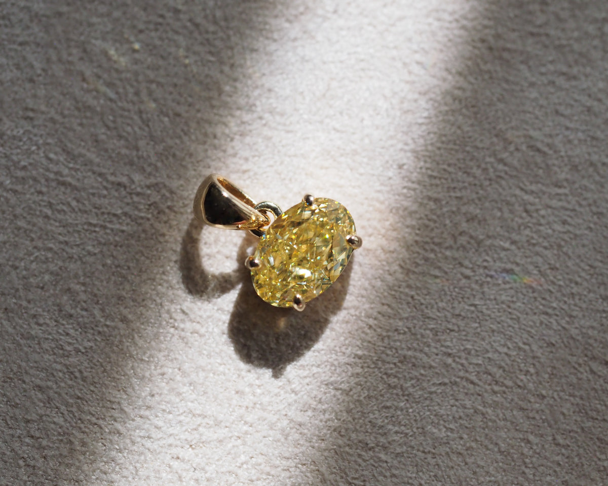 A yellow oval diamond pendant in a gold setting, resting on a textured light gray surface with dramatic diagonal shadows.