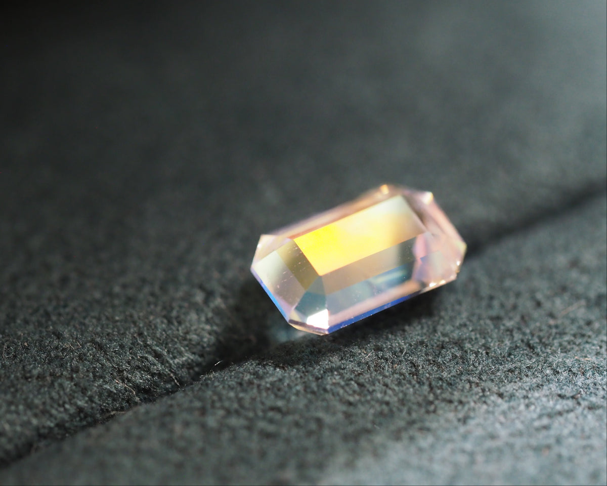 A single rectangular faceted gemstone with an aurora borealis iridescent finish, resting on a textured dark gray surface.