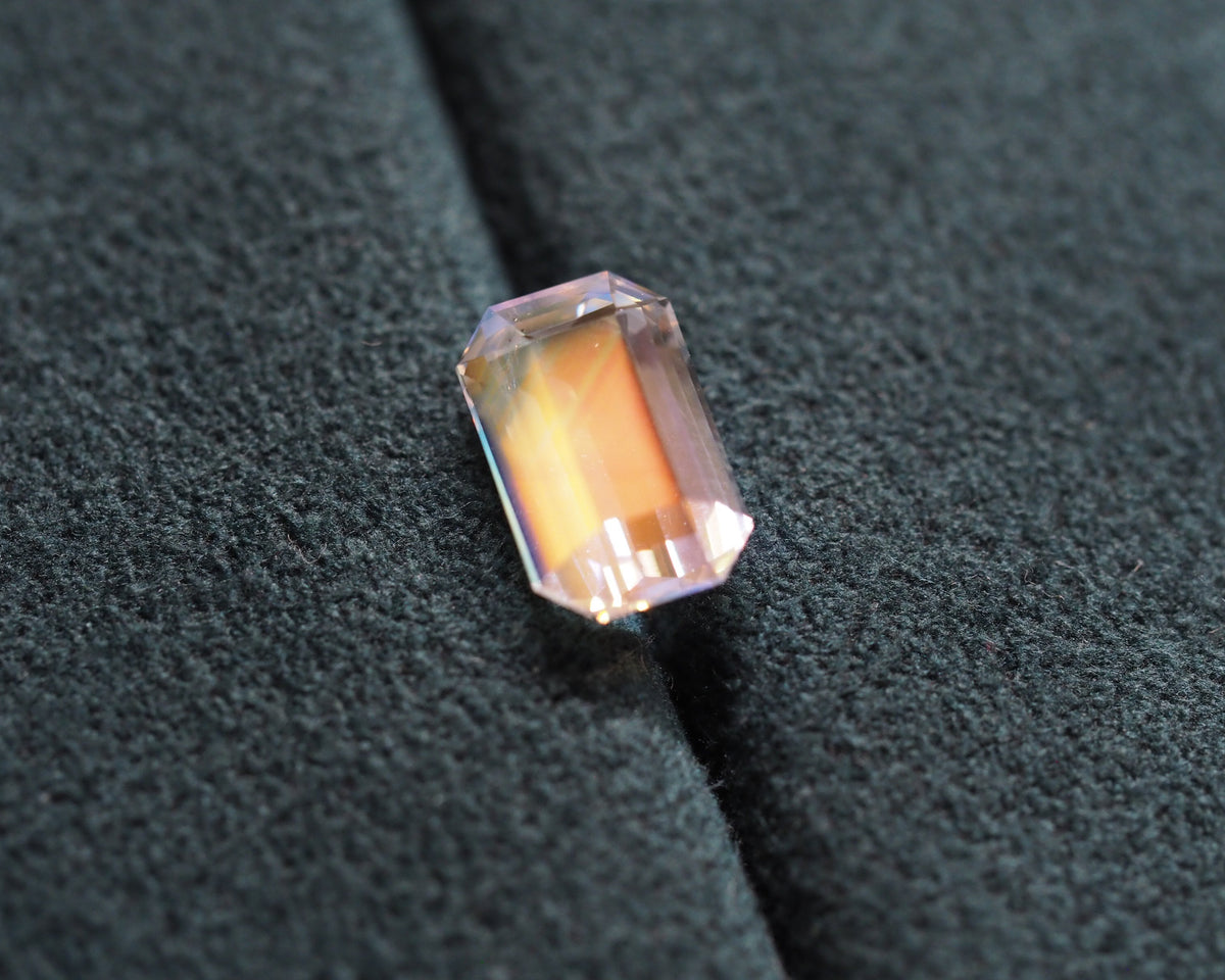 A rectangular emerald-cut pinkish-orange gemstone with a faceted surface, resting on a textured dark gray fabric background.