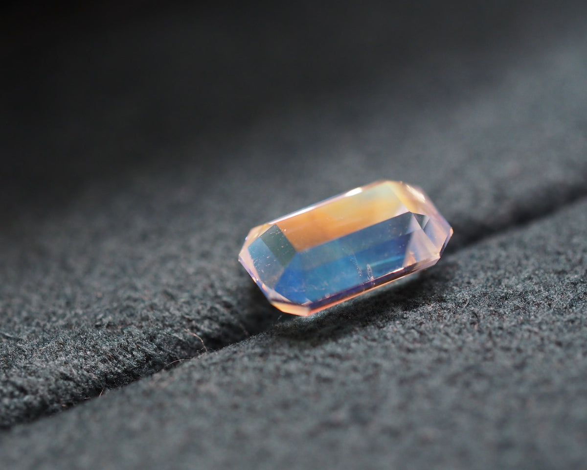 A rectangular faceted gemstone with a gradient color shift from warm peach-orange to deep blue, resting on a textured dark gray surface.