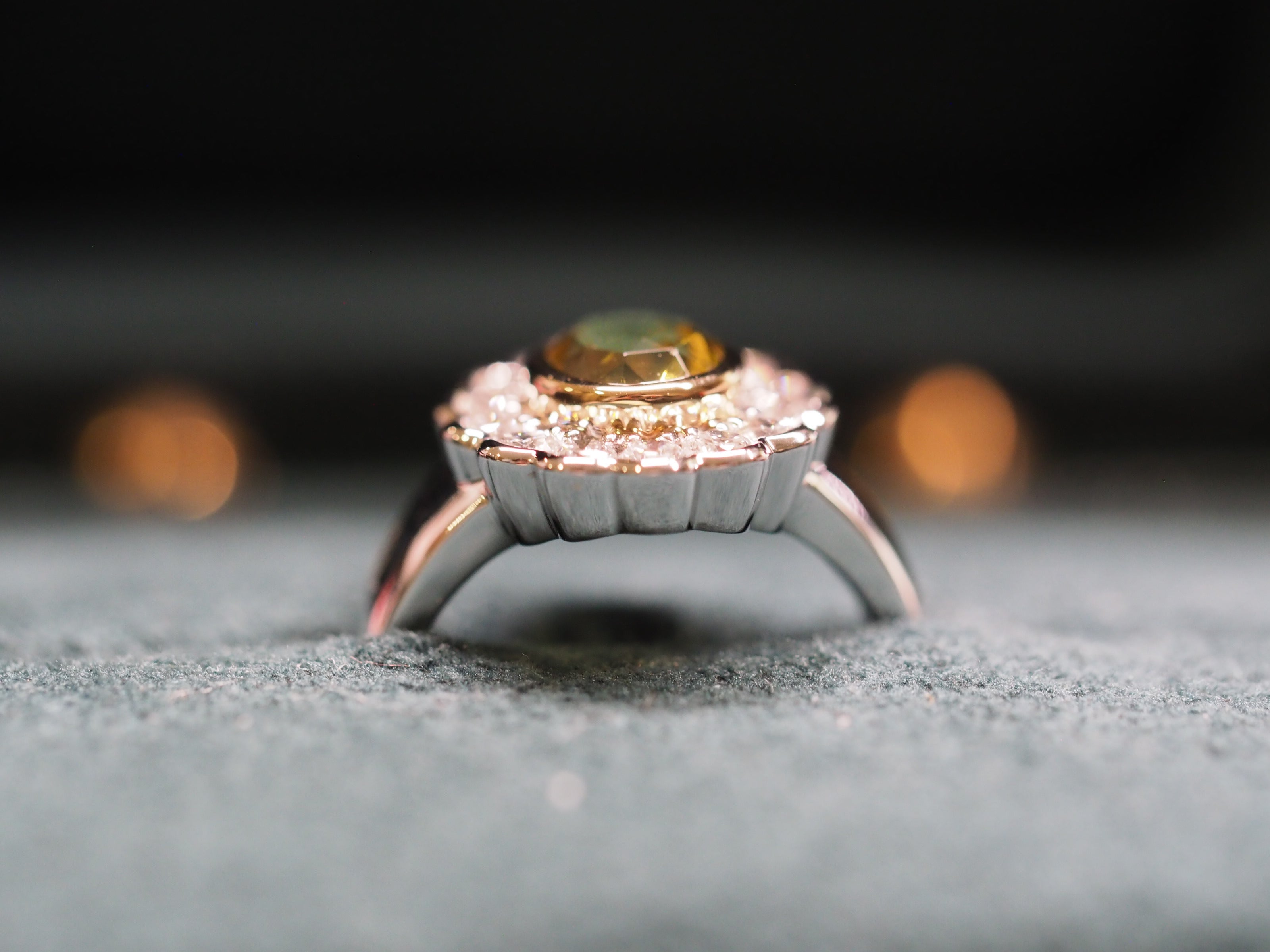 A close-up shot of a silver and yellow gold ring with a central lemon green chrysoberyl and small clear accent stones, resting on a textured gray surface.