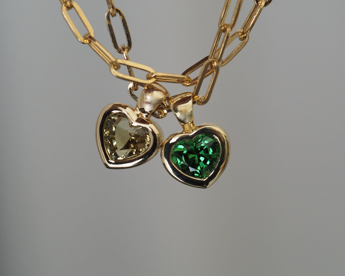 Gold chain necklace with two dangling heart charms, one with a clear faceted stone and one with a green faceted stone, both set in gold, shown against a plain gray background.