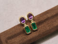 A pair of gold drop earrings with oval purple gemstones on top and rectangular green gemstones below, displayed on a brown textured stand against a light neutral background.