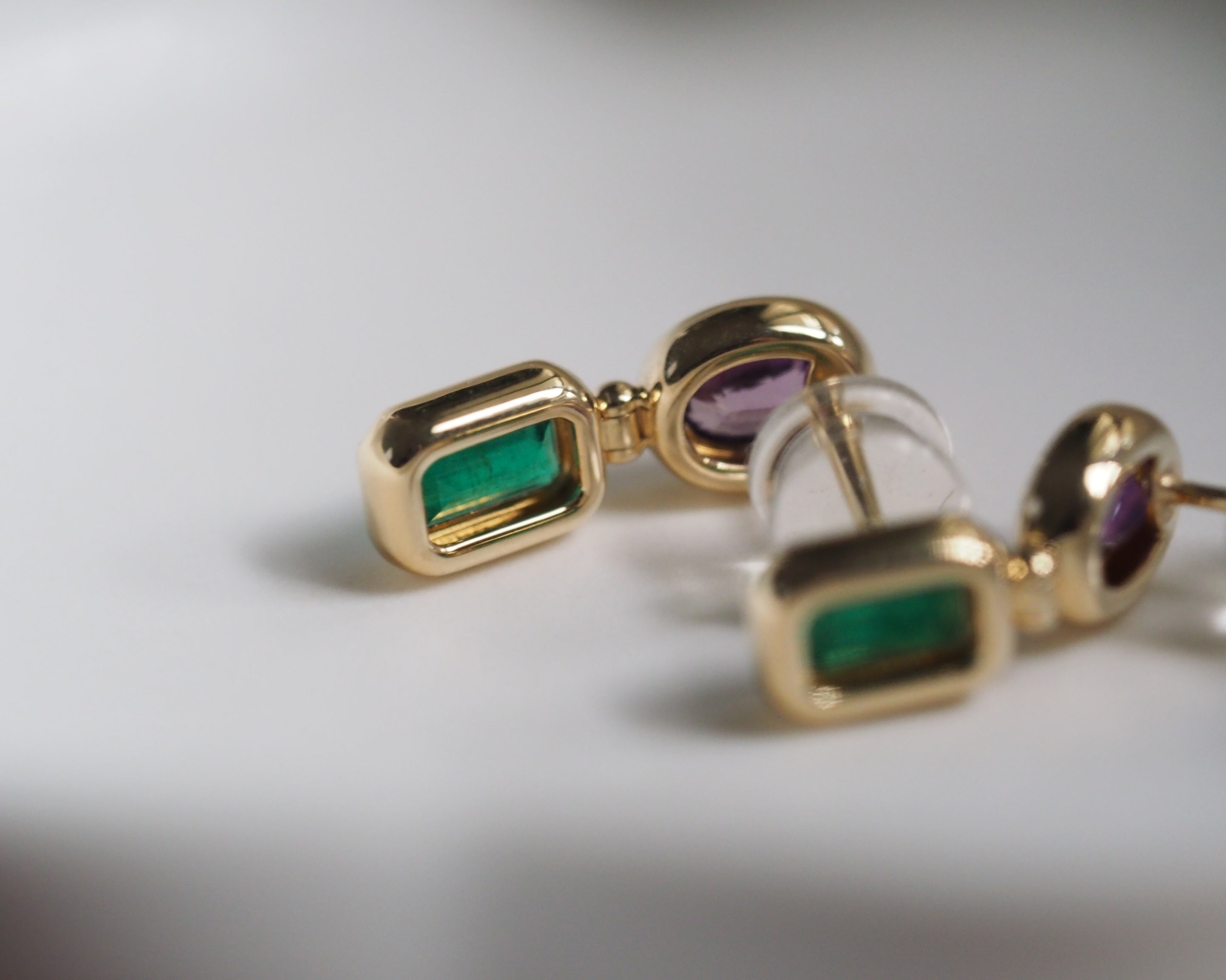 Gold-toned rectangular stud earrings with green emerald-cut stones and purple oval accent stones, shown on a white surface.