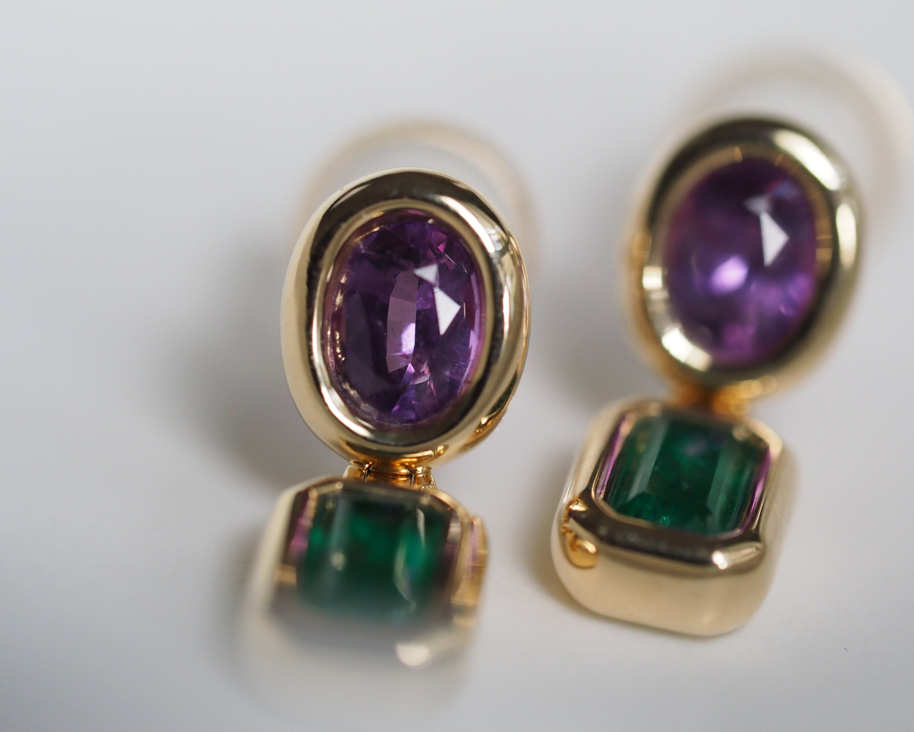 A pair of gold drop earrings featuring oval purple amethyst top stones and rectangular green emerald-cut bottom stones, shown on a clean white background.