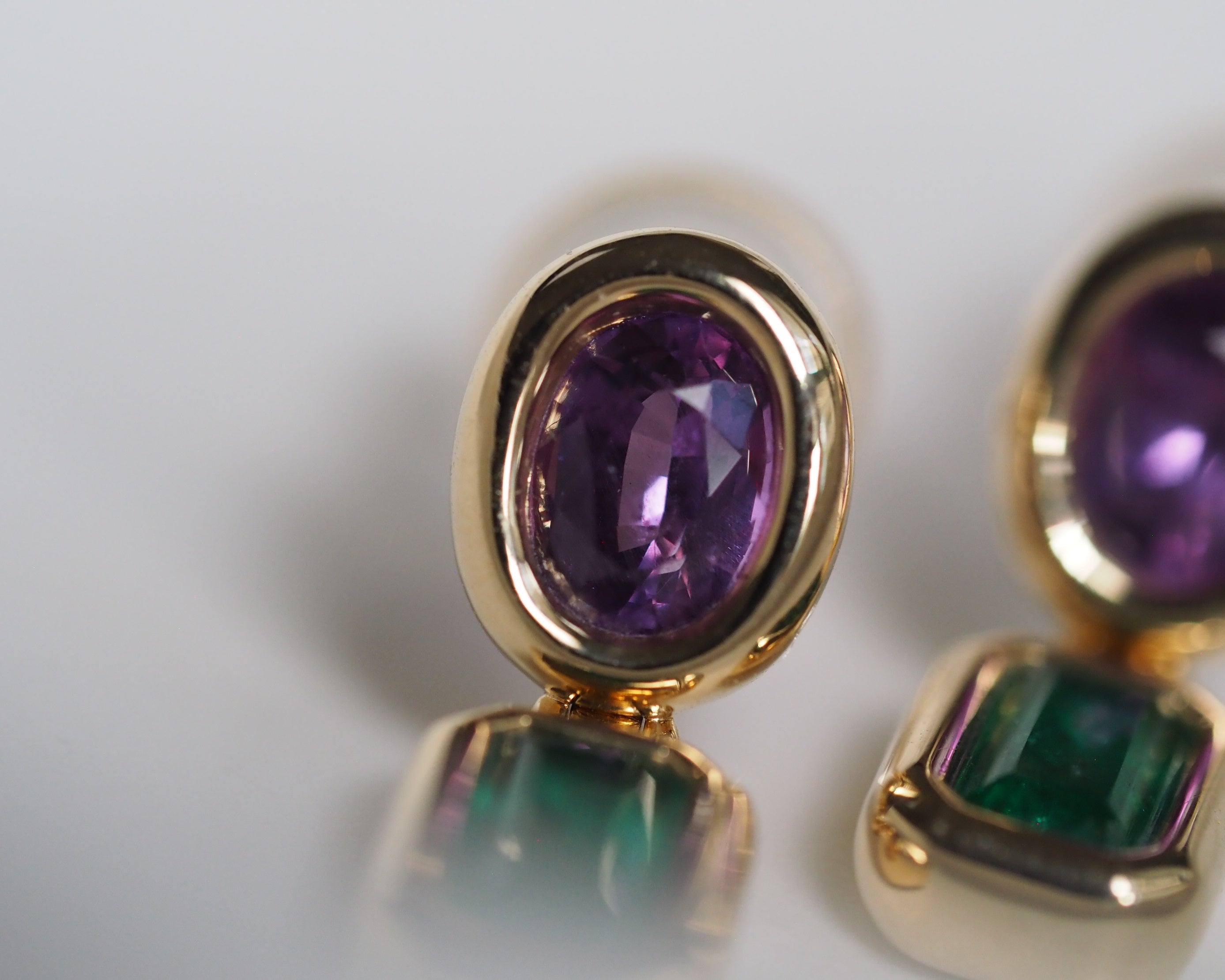 Gold-toned stud earrings with oval purple amethyst centers and rectangular green emerald-cut stones, shown in a close-up studio shot on a light background.