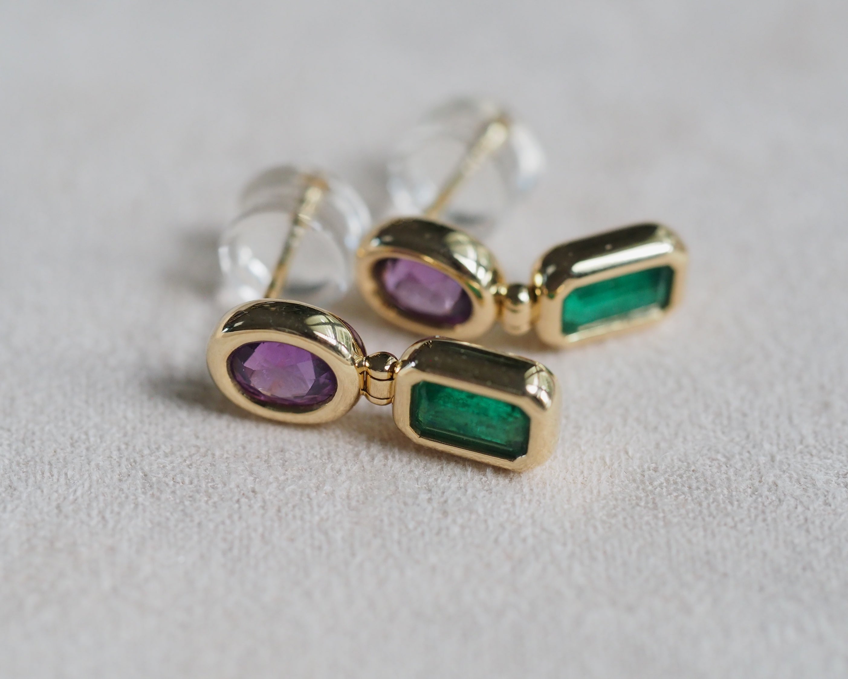 A pair of gold drop earrings with oval purple gemstones and rectangular green gemstones, shown on a textured light gray surface.