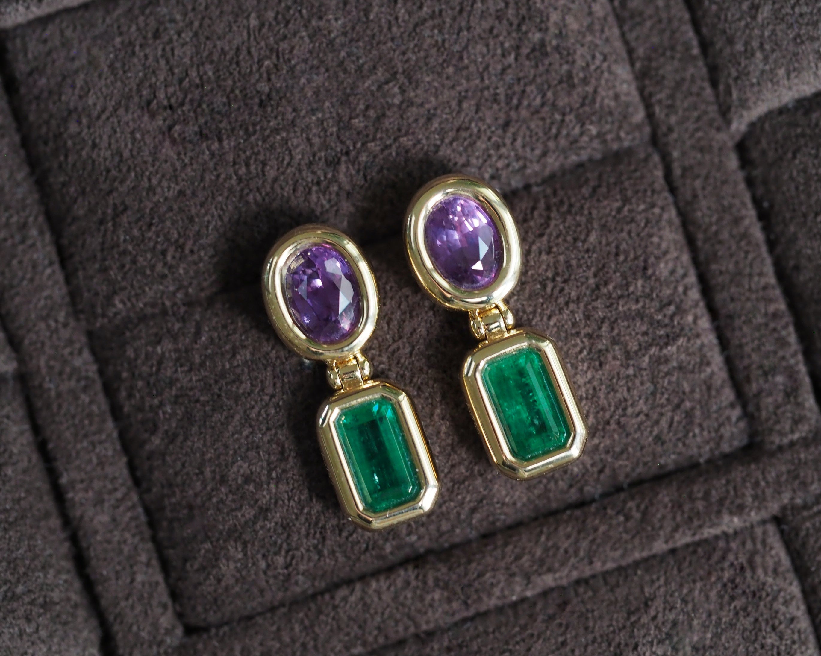 A pair of gold drop earrings featuring oval purple gemstones at the top and rectangular emerald-cut green gemstones below, displayed on a dark textured velvet surface.