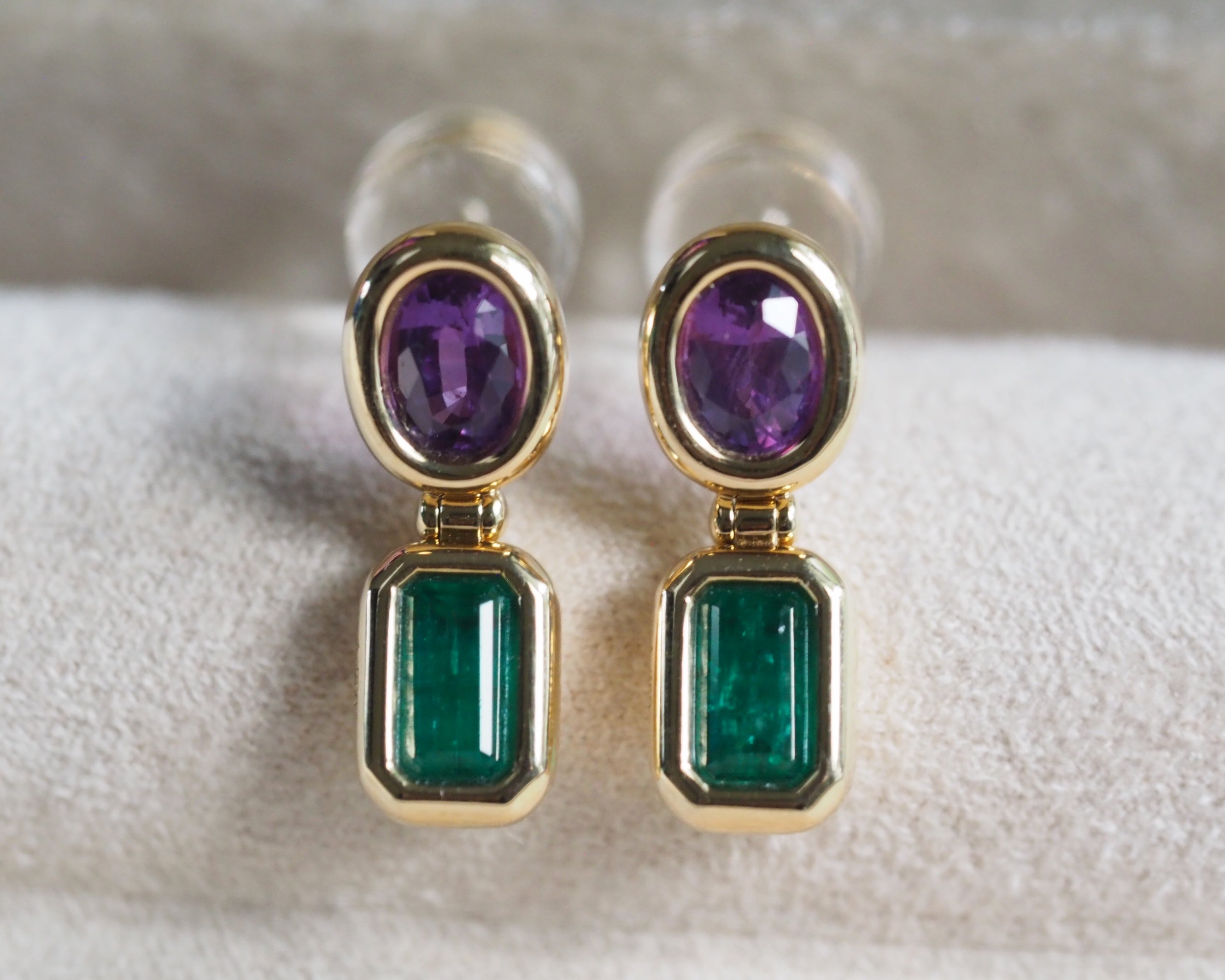 A pair of gold drop earrings featuring oval purple gemstones on top and rectangular green emerald-cut gemstones below, displayed on a soft white textured surface.